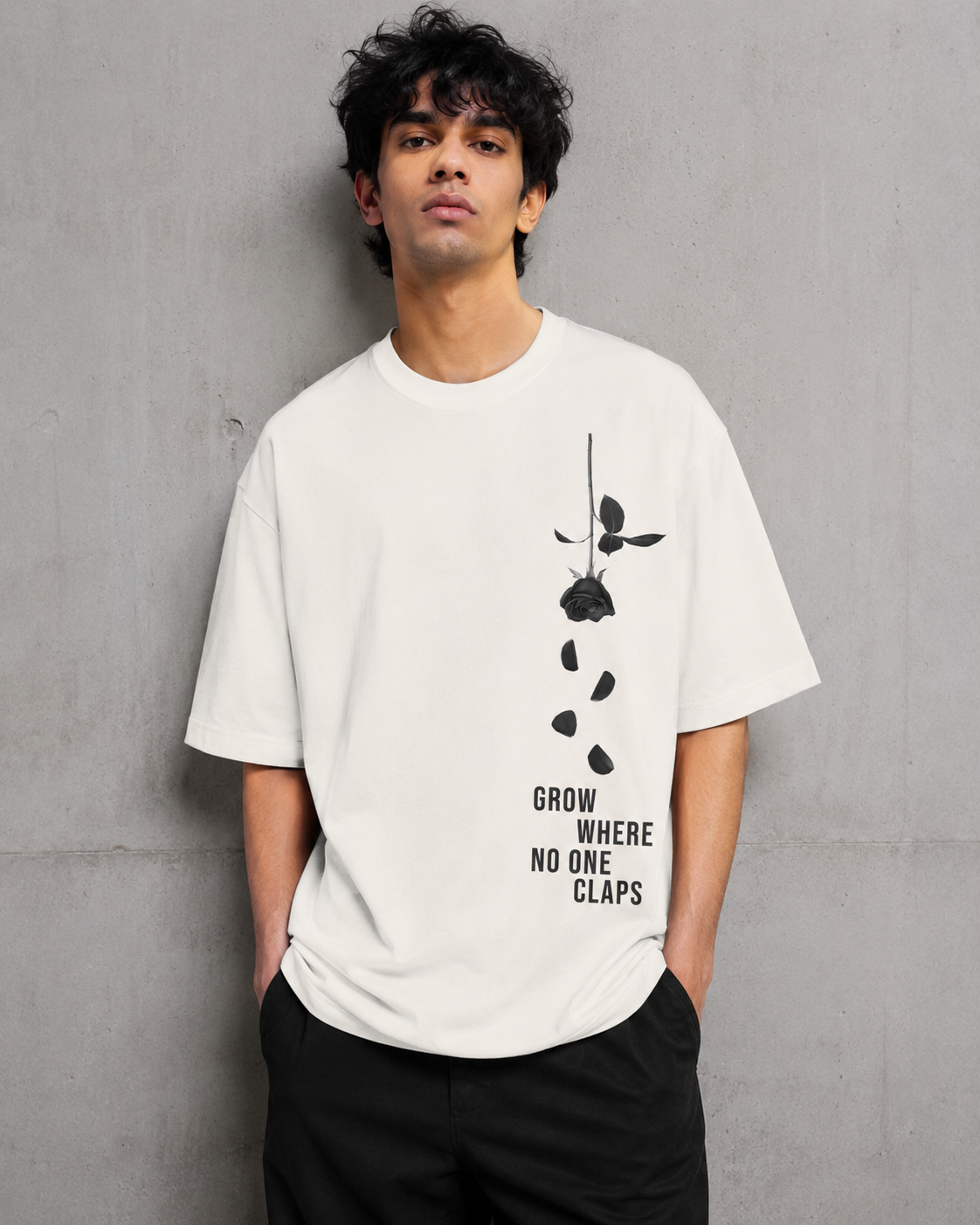A man wearing a white graphic t-shirt with a black rose design and the text 'GROW WHERE NO ONE CLAPS' on the front, standing against a textured gray concrete wall.