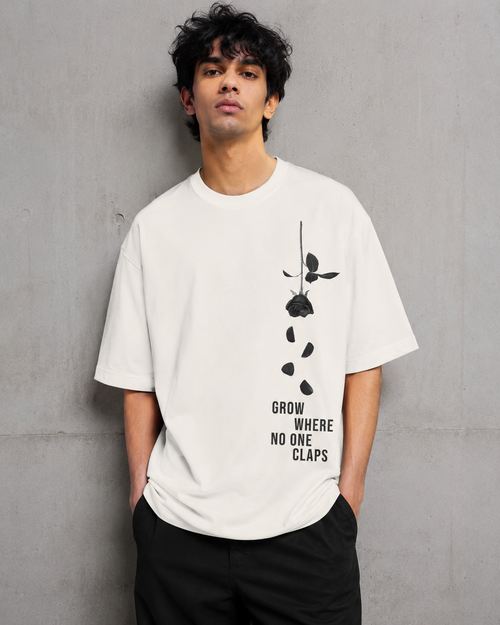 A man wearing a white graphic t-shirt with a black rose design and the text 'GROW WHERE NO ONE CLAPS' on the front, standing against a textured gray concrete wall.