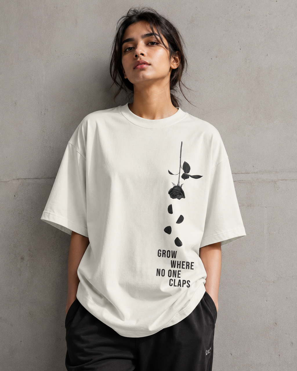 A woman wearing an oversized off-white t-shirt with a black rose graphic and the text 'GROW WHERE NO ONE CLAPS' on the front, paired with black sweatpants, standing against a textured gray concrete wall.