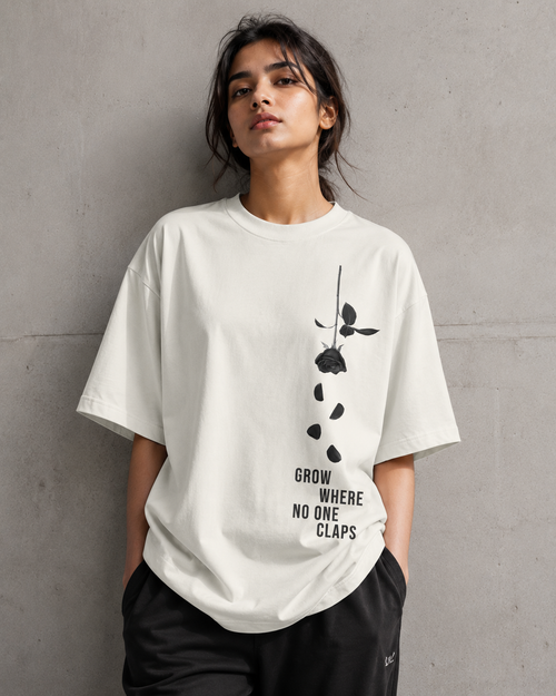 A woman wearing an oversized off-white t-shirt with a black rose graphic and the text 'GROW WHERE NO ONE CLAPS' on the front, paired with black sweatpants, standing against a textured gray concrete wall.