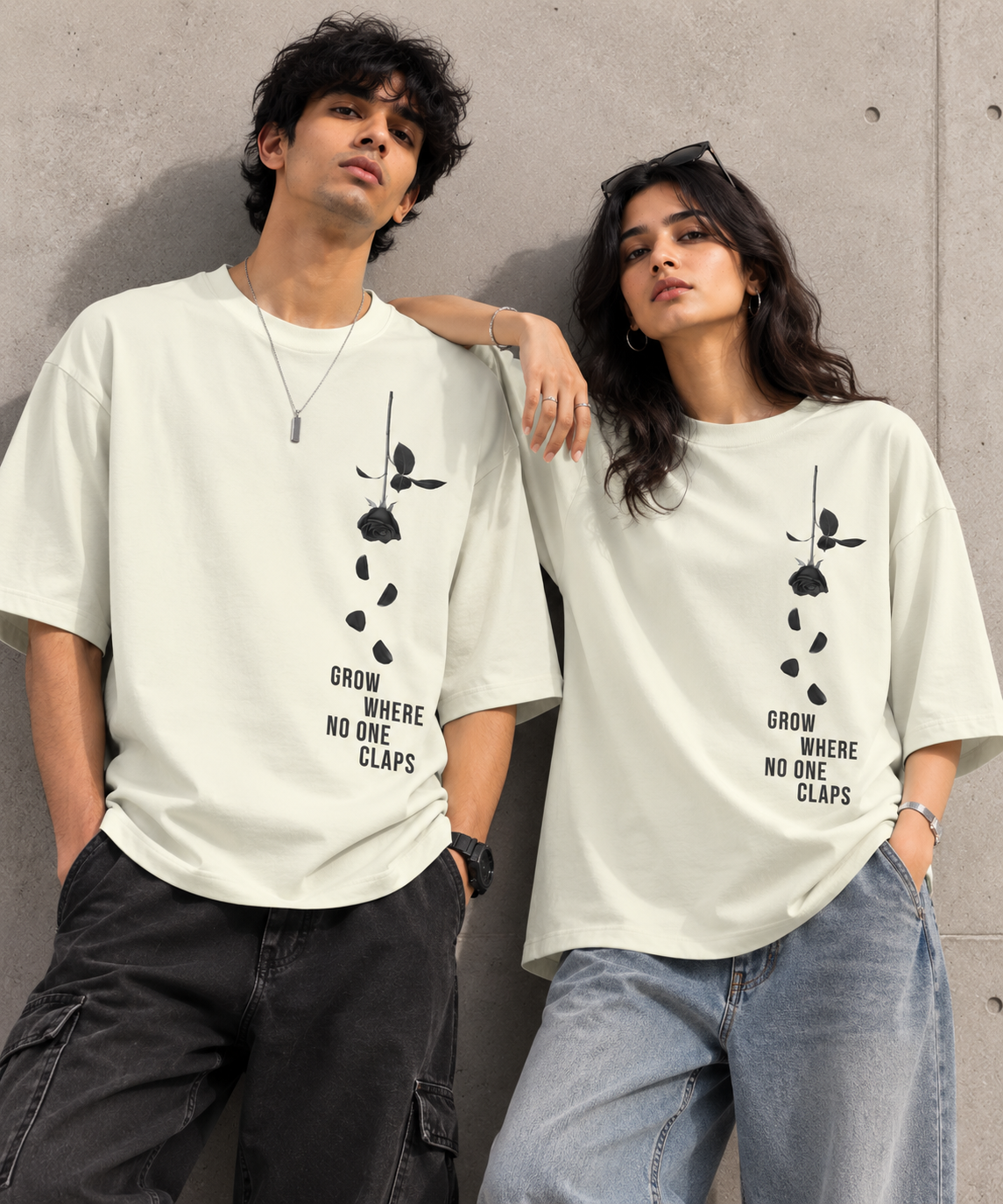 A man and a woman wearing matching off-white oversized t-shirts with a black rose graphic and the text 'GROW WHERE NO ONE CLAPS' printed on the front, posed against a concrete wall.